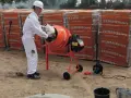 Budowlaniec w białym kombinezonie wsypuje składniki do betoniarki. W tle palety z cegłami. Klasy betonu czekają na użycie.