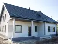 Nowy dom z szarą dachówką i jasną elewacją, ozdobioną subtelnymi, poziomymi pasami. Trwają prace przy malowaniu elewacji, tworząc nowoczesny wygląd.