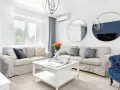 Jasny salon w stylu hampton z beżowymi sofami, niebieskimi akcentami i okrągłymi lustrami.