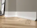 Białe, nowoczesne cokoły tworzą elegancką linię przy drewnianej podłodze w jodełkę. W tle widać fragment ściany i dekoracje.