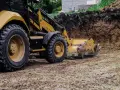 Żółta koparko-ładowarka pracuje przy niwelacji terenu, usuwając ziemię z wykopu.