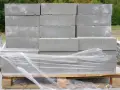 Stos szarych bloczków betonowych na palecie, owiniętych folią. Wymiary bloczków są standardowe, idealne do budowy.