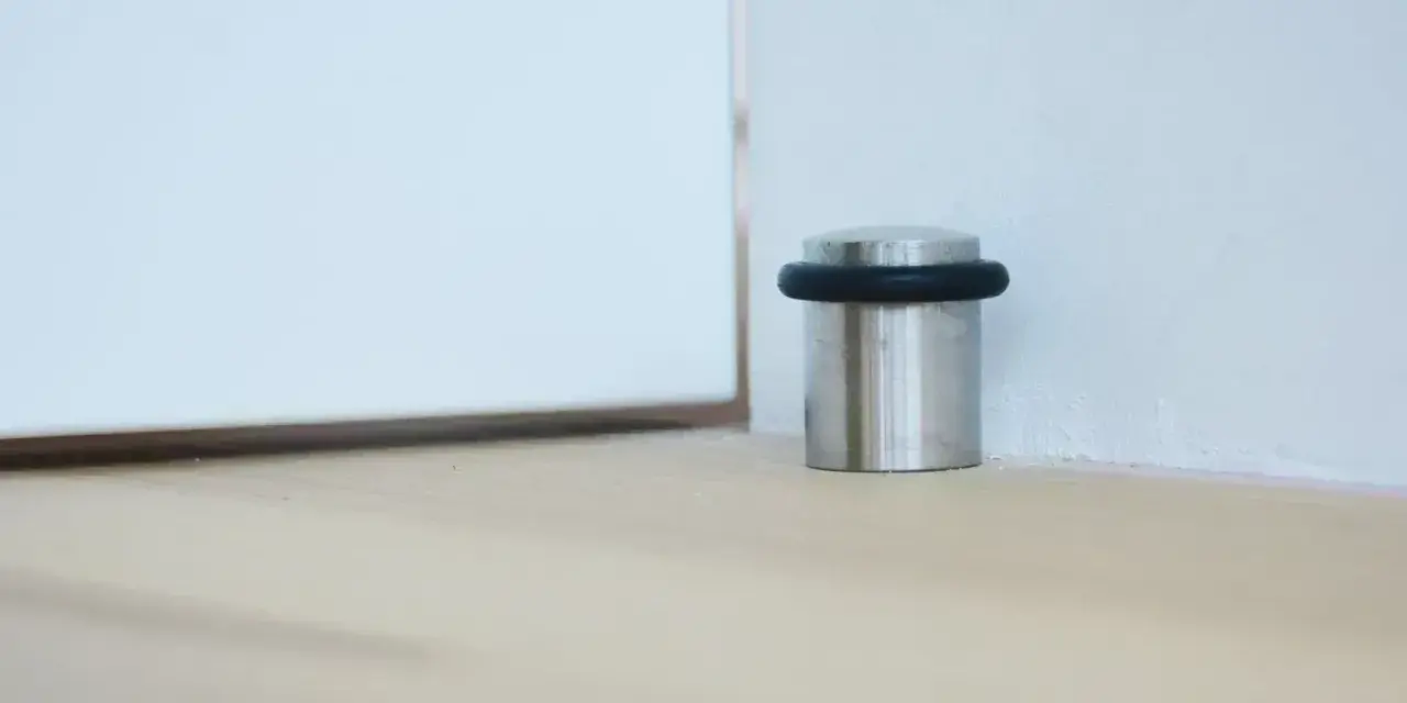 Metalowy odbojnik z czarną gumką przy ścianie, chroniący drzwi przed uderzeniem.