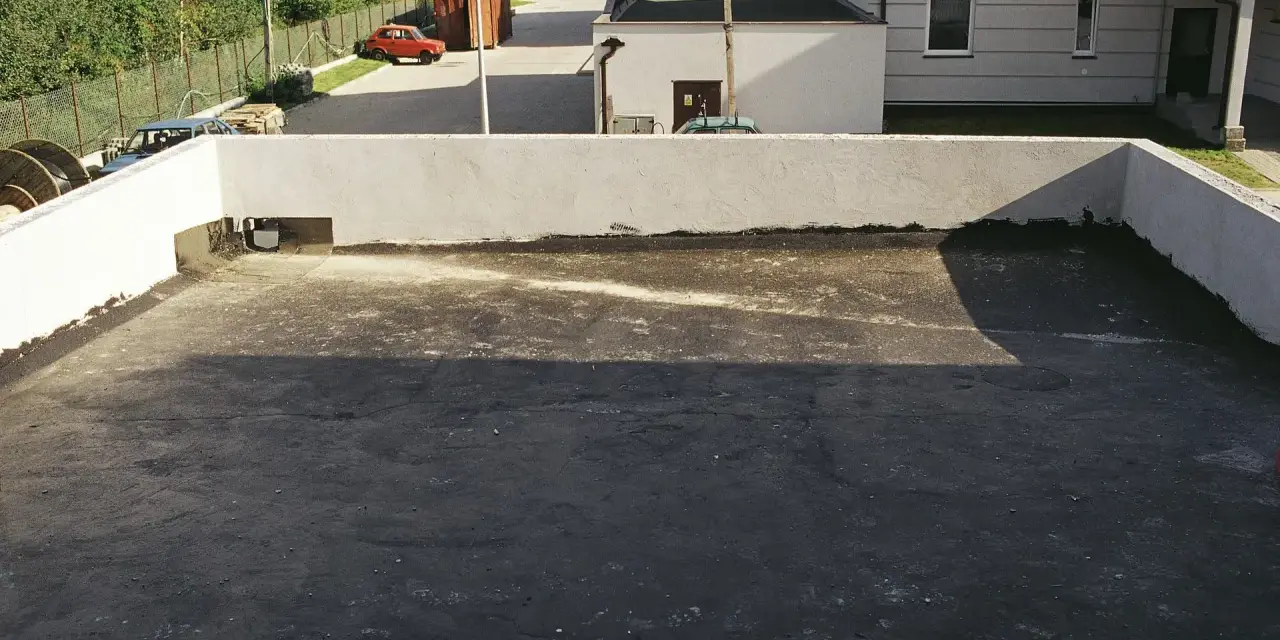 Płaski dach z papą, widoczny rzygacz odprowadzający wodę. W tle budynki i czerwony samochód.