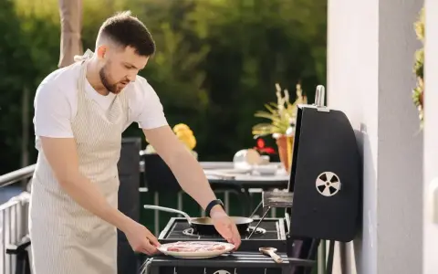 Mężczyzna w fartuchu przygotowuje mięso na grillu na balkonie. Czy można robić grilla na balkonie? Tak, jeśli jest bezpiecznie!