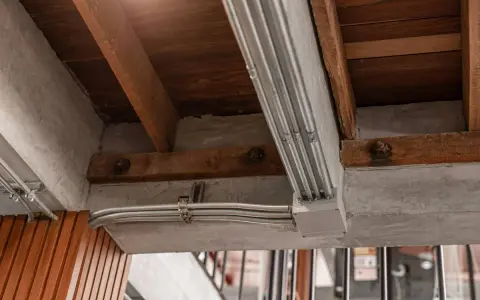 Metalowe rury elektryczne biegną wzdłuż betonowego podciągu, pod drewnianymi belkami stropowymi.