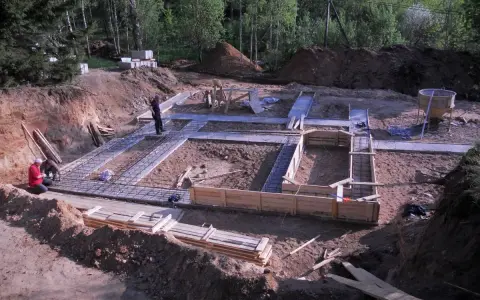 Budowa fundamentów domu. Wykop, zbrojenie i szalunki. W tle las, pracują dwaj chudziaki.