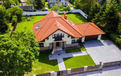 Piękny dom 200m2 z pomarańczowym dachem, otoczony zielonym ogrodem i drzewami. Podjazd z kostki brukowej prowadzi do garażu.