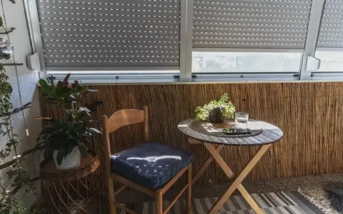 Przytulna loggia z bambusową ścianką, drewnianym krzesłem i stolikiem. Na stoliku szklanka wody i telefon.