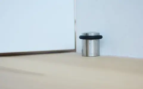 Metalowy odbojnik z czarną gumką przy ścianie, chroniący drzwi przed uderzeniem.