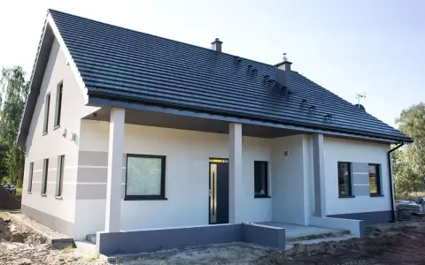 Nowy dom z szarą dachówką i jasną elewacją, ozdobioną subtelnymi, poziomymi pasami. Trwają prace przy malowaniu elewacji, tworząc nowoczesny wygląd.
