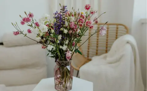Wazon z kolorowymi kwiatami ciętymi, wśród nich różowe dzwonki i białe margaretki, stoi na stoliku obok plecionej wiklinowej fotel.