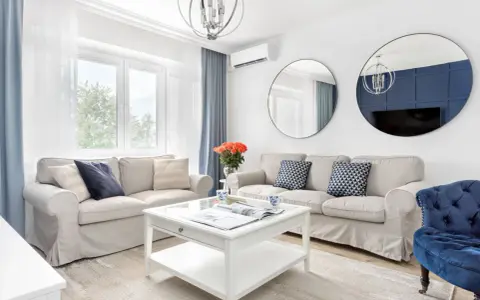 Jasny salon w stylu hampton z beżowymi sofami, niebieskimi akcentami i okrągłymi lustrami.