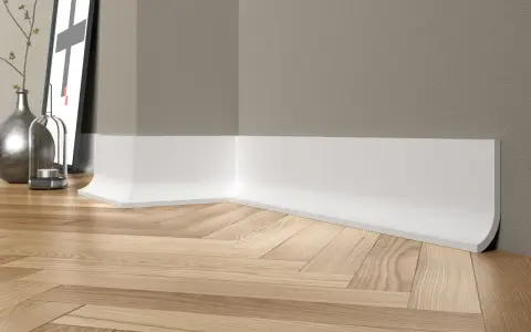 Białe, nowoczesne cokoły tworzą elegancką linię przy drewnianej podłodze w jodełkę. W tle widać fragment ściany i dekoracje.