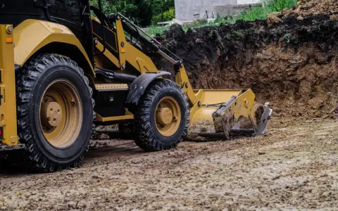 Żółta koparko-ładowarka pracuje przy niwelacji terenu, usuwając ziemię z wykopu.