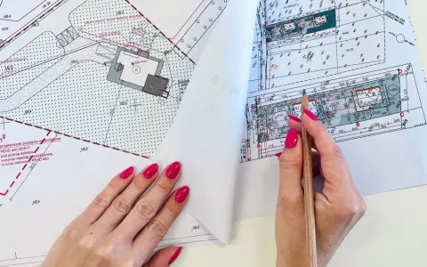 Ręka z różowymi paznokciami wskazuje ołówek na mapie do celów projektowych, gdzie widoczne są plany budowy i przyłączy.
