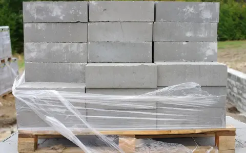 Stos szarych bloczków betonowych na palecie, owiniętych folią. Wymiary bloczków są standardowe, idealne do budowy.