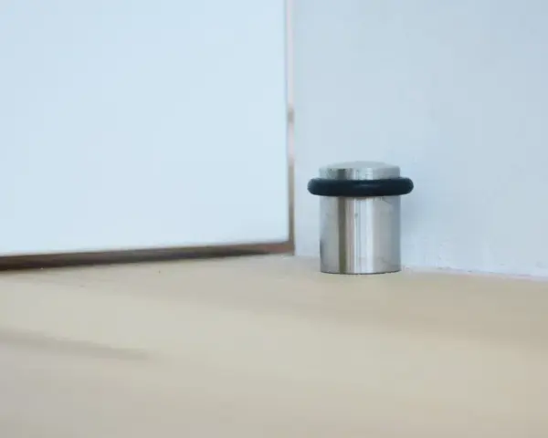 Metalowy odbojnik z czarną gumką przy ścianie, chroniący drzwi przed uderzeniem.