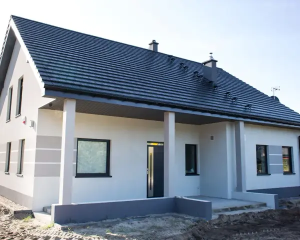 Nowy dom z szarą dachówką i jasną elewacją, ozdobioną subtelnymi, poziomymi pasami. Trwają prace przy malowaniu elewacji, tworząc nowoczesny wygląd.