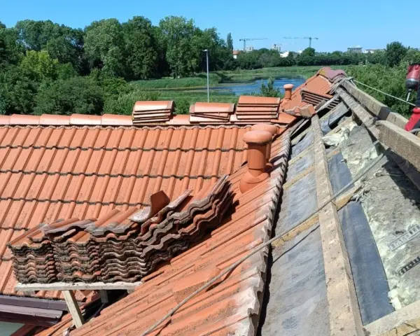Trwa wymiana dachu. Stare dachówki są ułożone w stosy, a pod nimi widać izolację i drewniane elementy konstrukcji.