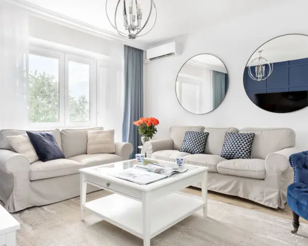 Jasny salon w stylu hampton z beżowymi sofami, niebieskimi akcentami i okrągłymi lustrami.