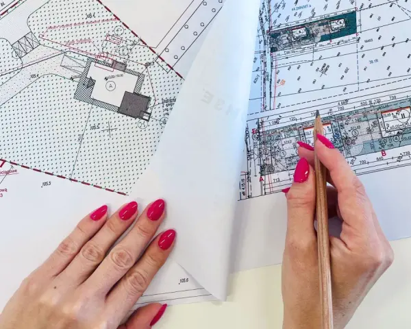 Ręka z różowymi paznokciami wskazuje ołówek na mapie do celów projektowych, gdzie widoczne są plany budowy i przyłączy.