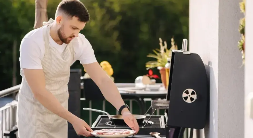 Mężczyzna w fartuchu przygotowuje mięso na grillu na balkonie. Czy można robić grilla na balkonie? Tak, jeśli jest bezpiecznie!