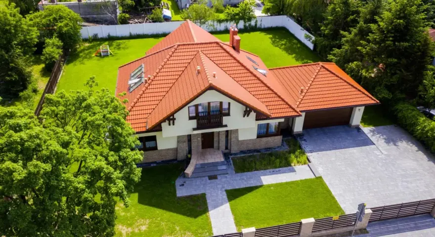 Piękny dom 200m2 z pomarańczowym dachem, otoczony zielonym ogrodem i drzewami. Podjazd z kostki brukowej prowadzi do garażu.