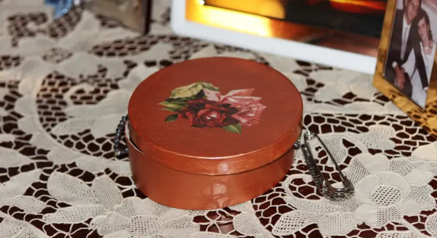 Okrągłe pudełko ozdobione techniką decoupage z motywem róż. Obok broszka i ramka ze zdjęciem.