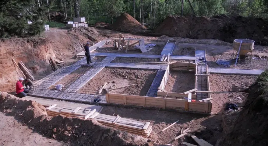 Budowa fundamentów: zbrojenie i szalunki czekają na chudy beton. W tle las i plac budowy.