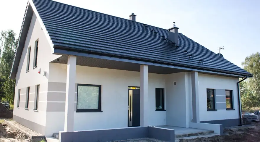 Nowy dom z szarą dachówką i jasną elewacją, ozdobioną subtelnymi, poziomymi pasami. Trwają prace przy malowaniu elewacji, tworząc nowoczesny wygląd.