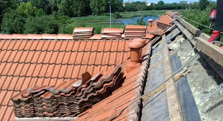 Trwa wymiana dachu. Stare dachówki są ułożone w stosy, a pod nimi widać izolację i drewniane elementy konstrukcji.