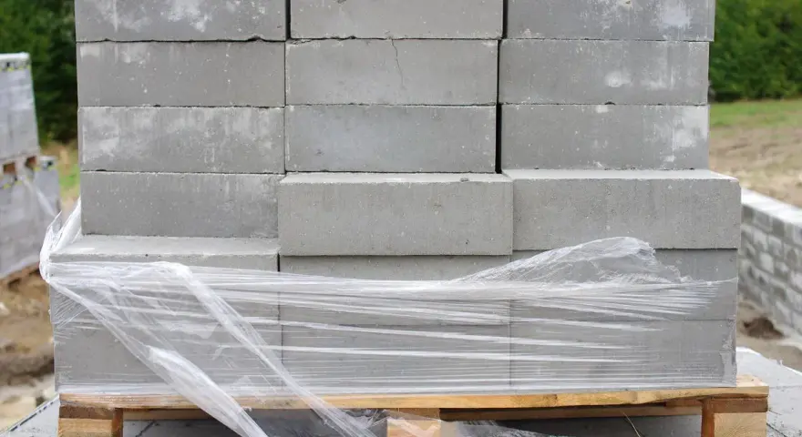 Stos szarych bloczków betonowych na palecie, owiniętych folią. Wymiary bloczków są standardowe, idealne do budowy.