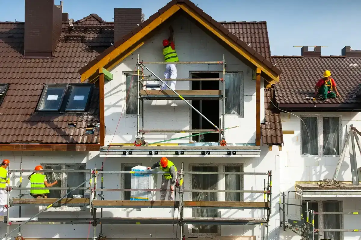 Pracownicy na rusztowaniach prowadzą termomodernizację budynku, ocieplając ściany i dach.