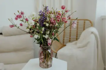 Wazon z kolorowymi kwiatami ciętymi, wśród nich różowe dzwonki i białe margaretki, stoi na stoliku obok plecionej wiklinowej fotel.